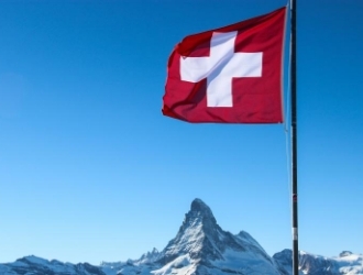 An image of the flag of Switzerland flying on a flagpole with mountains in the background